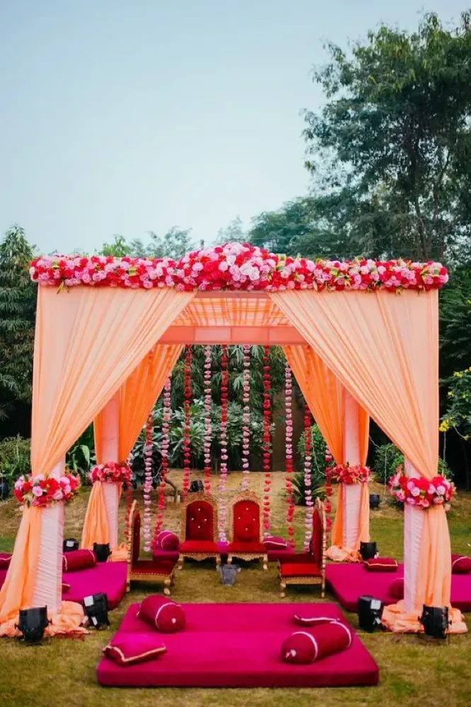 Outdoor wedding mandap with peach drapes, a pink and red rose canopy, and traditional ceremonial seating