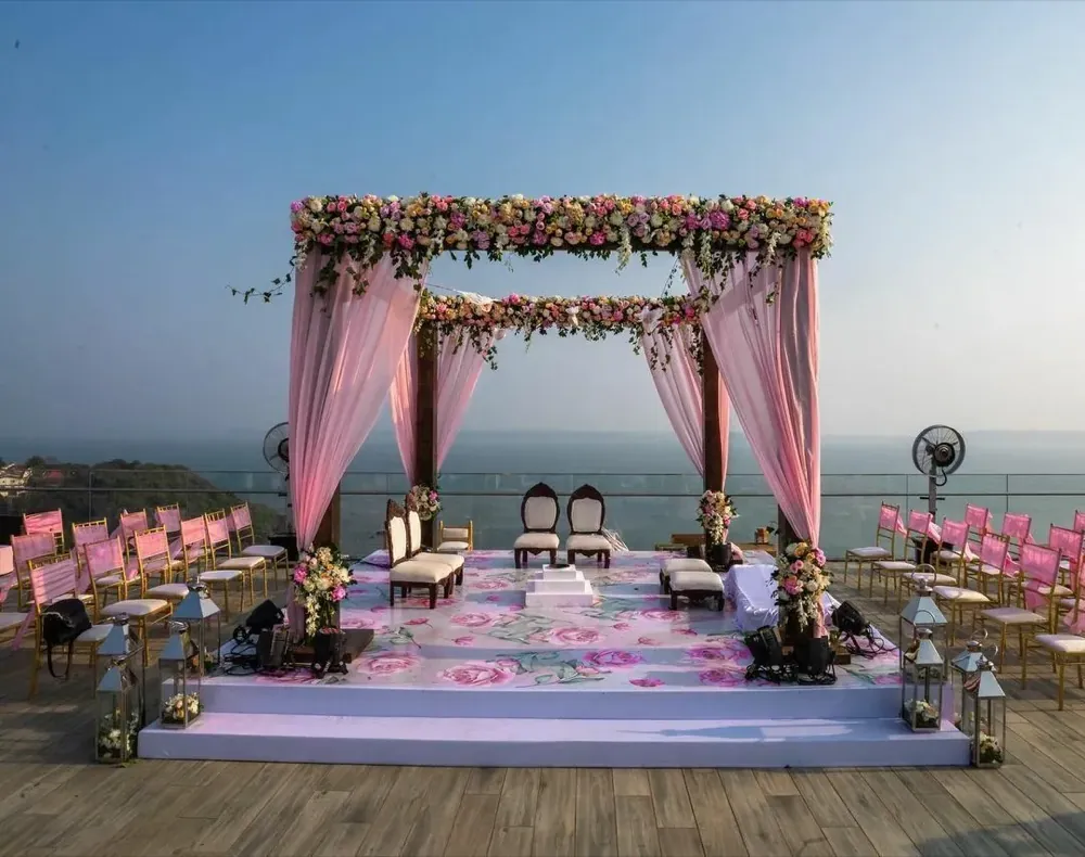 Rooftop wedding mandap with pastel pink drapes, a floral canopy, and a rose-print stage overlooking the sea