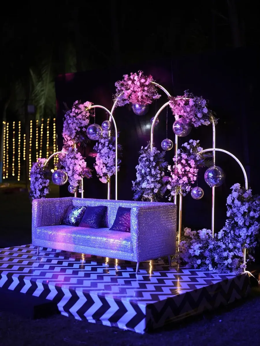Evening sangeet stage with a sequinned sofa, pink-lit floral arches, and a black-and-white chevron floor