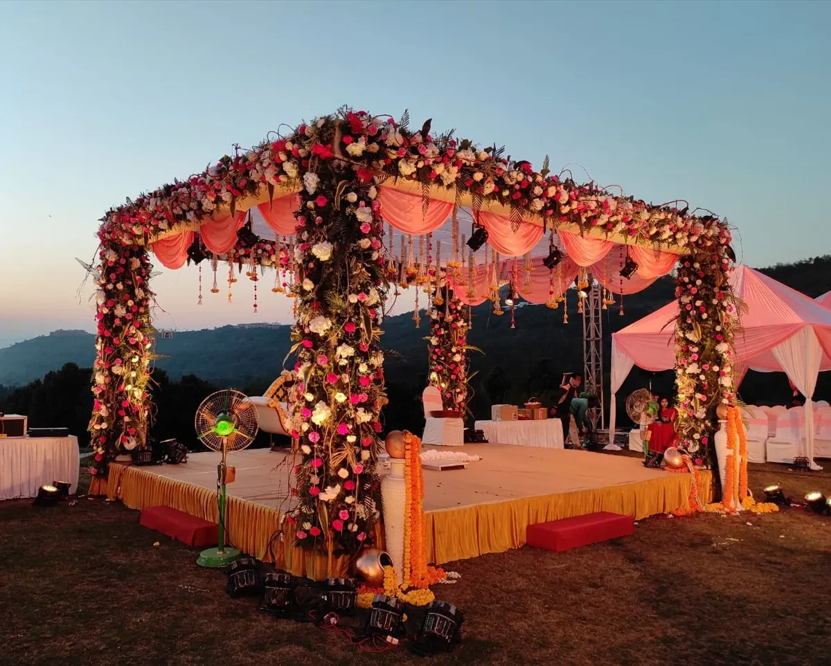 Open-air hilltop wedding mandap at dusk with rose-wrapped pillars, a peach canopy, and marigold garlands